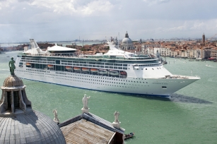 The image shows a large white cruise ship, Grandeur of the Seas, sailing through a wide greenish-blue waterway, likely near a historic coastal city. In the background, you can see the iconic skyline of Venice, Italy, with its domed churches, bell towers, and red-roofed buildings stretching across the horizon. 